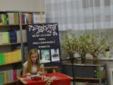 Uczennica gimnazjum siedzi przy stoliku i czyta książkę. Obok na parapecie stoją trzy wazony z kwiatami.