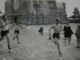 Czarno-białe zdjęcie, trzech chłopców w krótkich spodenkach biegnących po piaszczystej bieżni. W tle budynek szkoły.