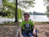 Chłopiec w zielonej koszulce siedzi w wózku inwalidzkim. W tle tafla jeziora