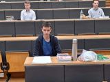 W auli wykładowej przy długim stole siedzi uczeń liceum w niebieskiej koszulce i czarnej bluzie i rozwiązuje test.
