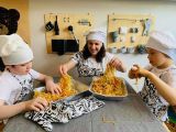Dwoje uczniów szkoły podstawowej i uśmiechnięta kobieta w czapkach kucharskich trzymają w dłoniach długi makaron spaghetti