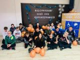 Grupa uczniów szkoły podstawowej siedzi na podłodze trzymając w dłoniach pomarańczowe balony