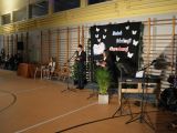 Sala gimnastyczna. Ubrany w garnitur nastolatek stoi przed mikrofenem. W tle dekoracje na drabinkach gimastycznych.
