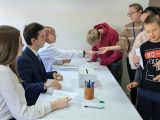 Grupa uczniów pobiera karty do głosowania. Po lewej uczniowie w strojach galowych