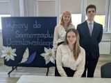 Troje uczniów w strojach galowych. Po lewej granatowa tablica z napisem Wybory do Samorządu Uczniowskiego