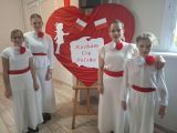 Cztery uczennice w białych sukniach śpieea przez mikrofon. Między nimi duże czerwone serce z napisem Kocham Cię Polsko...