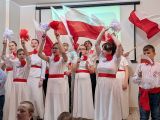 Grupa uczniów w biało - czerwonych strojach z rękami uniesionymi do góry. Z tyłu grupa ucziów machająca flagami Polski