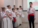 Mężczyzna w białej koszuli przepasany czerwoną szarfą śpiewa przez mikrofon. Po lewej grupa uczniów w białych strojach z...