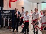 Mężczyzna w granatowym garniturze mówi przez mikrofon. Obok grupa uczniów w strojach galowych. Po lewej tablica z napisem...