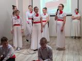 Uczennica w białej sukni śpiewa trzymajać mikrofon. Z przodu dwóch chłopców siedzących po turecku. Obok grupa młodzieży w...