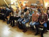 Duża grupa uczniów liceum siedzi na krzesłach w kilku rzędach w sali szkolnej. Przy ścianach wiszą stare zdjęcia.