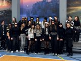 Grupa nastolatków stoi pośrodku sali gimnastycznej. Pośród nich stoi kobieta w okularach. Wszyscy mają na sobie część...