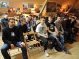 Duża grupa uczniów liceum i mężczyzna siedzą na krzesłach w kilku rzędach w sali szkolnej. Przy ścianach wiszą stare zdjęcia.