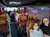 Uczniowie i kobieta w okularach siedzą we wnętrzu autobusu. Autobus z pomarańczowo-żółtymi oparciami siedzeń