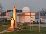 Biały budenek z kopułą. Przed budynkiem na trawniku granitowy pomnik mężczyzny.