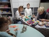 Czworo uczniów przy owalnych stolikach. Na stolikach przybory malarskie. W tle biały regał z szarymi szufladam