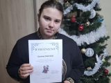 Uczennica w ciemnej bluzie z dyplomem w dłoni. Za nią przystrojona sztuczna choinka