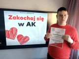 Uczeń w czerwonej bluzie stoi z dyplomem w ręku. W tle napis Zakochaj się w AK wyświetlany na dużym ekranie tablicy...