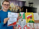 Uczeń w niebieskiej bluzie i w okularach pokazuje palcem pracę plastyczną o tematyce wojennej