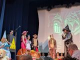 Mężczyzna z brodą w czarnym stroju i wysokim kapeluszu mówi przez mikrofon. Obok grupa uczniów w bajkowych, kolorowych...