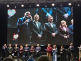 Kilkoro ludzi na ekranie. Poniżej szereg osób na scenie.
