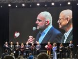 Dwóch mężczyzn na ekranie poniżej kilkanaście osób w szeregu na scenie