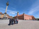 Grupa osób na dużym placu. W tle czerwony budynek z wieżą zakończoną kopułą i wysoka kolumna z postacią króla na szczycie