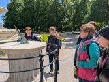 Kobieta w kolorowej kurtce i czworo uczniów przed zegarem słonecznym w parku
