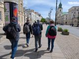 Pięcioosobowa grupa spaceruje szeroką ulicą. Po prawej i lewej stronie wysokie budynki