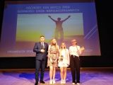 Dwie uśmiechnięte uczennice na scenie. Po lewej mężczyzna w szarym garniturze. Po prawej kobieta z warkoczem w białej bluzce