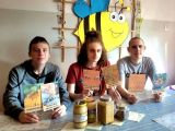 Trzech chłopców trzyma w dłoniach zdjecia pszczół, przed nimi produkty pszczele, w tle pszczoła z papieru.