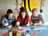 Trzech chłopców układa puzzle, przed nimi stoją produkty pszczele, w tle pszczoła z papieu