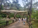 Trzech chłopców spaceruje ścieżką wśród zieleni. Przed nimi model wielkiego dinozaura z długą szyją
