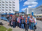 Grupa uczniów i dorosłych przed tramwajem kolejką. W tle wysokie białe budynki