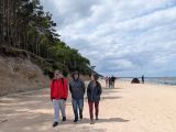 Troje uśmiechniętych uczniów w kurtkach i kapturach spaceruje po słonecznej, szerokiej plaży