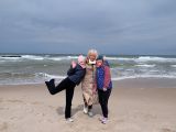 Uśmiechnięta kobietaz jasnymi włosami stoi na wietrznej plaży. Obok niej dwie dziewczynki ubrane w kurtki i czapki