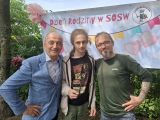 Dwóch uśmiechniętych mężczyzn i wysoki chłopak stojący między nimi. Mężczyzna po lewej w niebieskiej marynarce