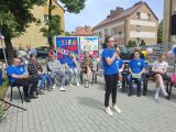 Dziewczyna w niebieskim t-shircie i okularach śpiewa przez mikrofon. Za nią grupa ludzi siedząca na krzesłach