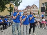 Trzy dziewczyny w niebieskich t-shirtach śpiewają przez mikrofony. W tle grupa uczniów siedząca na krzesłach