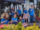 Uczeń w niebieskim t-shircie śpiewa przez mikrofon stojąc na scenie plenerowej. Oboch sziedzące dziewczynki
