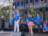 Uczniowie w niebieskich t-shirtach na scenie. Z przodu dziewczyna z długimi włosami śpiewająca przez mikrofon