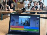 Na pierwszym planie laptop. W tle uczniowie w ławkach