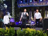 Dwaj mężczyźni na scenie. Na pierwszym planie zielone krzewy