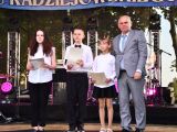 Scena . Troje dzieci w galowych strojach. Obok Mężczyzna w szarym garniturze