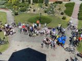 Na dziedzińcu przed szkołą stoi duza grupa osób z rowerami. Wszyscy w kolorowych strojach.
