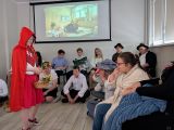 Dziewczynka w okularach w czerwonej pelerynie z koszyczkiem w dłoni. Obok grupa siedzących uczniów