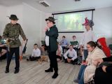 Dwóch chłopców w zielonych kapeluszach z piórkiem i strzelbami na ramionach. W tle grupa siedzących uczniów