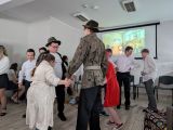 Dwóch uczniów w kapeluszach z piórkami i wojskowych strojach tańczy w kręgu trzymając za ręce dziewczynkę w szlafroku