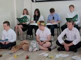 Dziewczyna w wianku z kolorowych kwiatów na głowie. Obok dwóch uczniów w strojach galowych siedzących po turecku