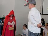 Dziewczynka w czerwonej pelerynie z kapturem. Obok chłopak w białej koszuli w czapce wilka na głowie